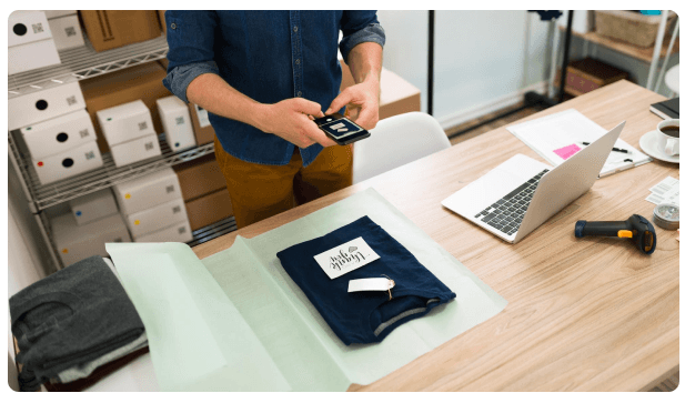 Man photographing a folded garment with a phone next to a laptop and stacked boxes