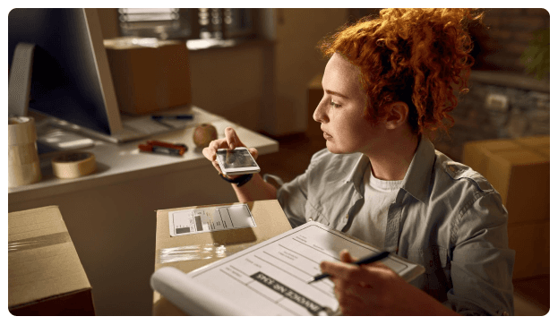 Woman with curly red hair scanning a package with a phone while holding a clipboard