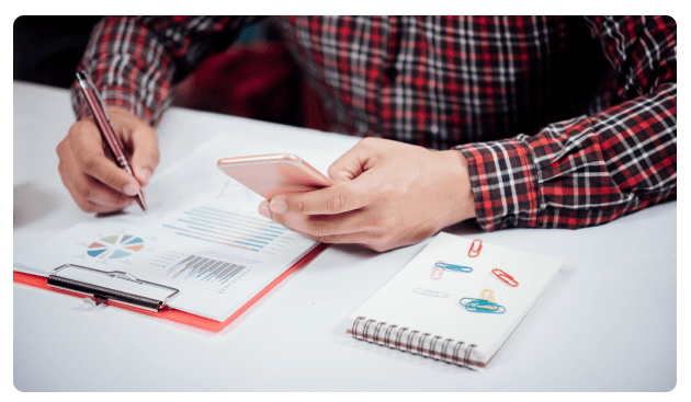Man in a plaid shirt writing on a report while holding a phone, with a notebook of paperclips nearby