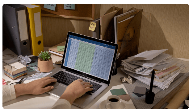 Hands typing on a laptop displaying a spreadsheet, with a cup of coffee and paint swatches on a desk