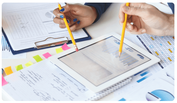 Two people pointing pencils at a tablet displaying financial charts
