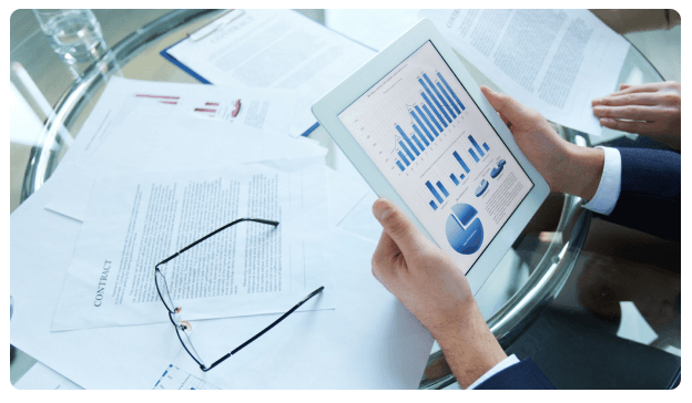Hand holding a tablet displaying bar and pie charts beside glasses and a contract