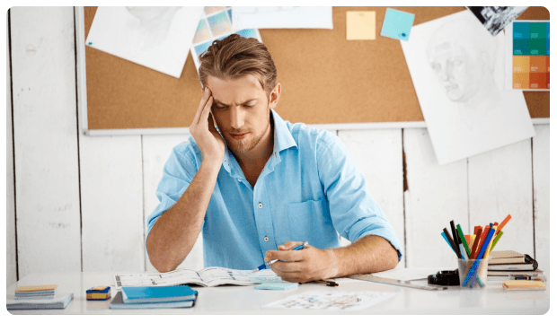 Man resting his head on his hand at a desk in front of a corkboard